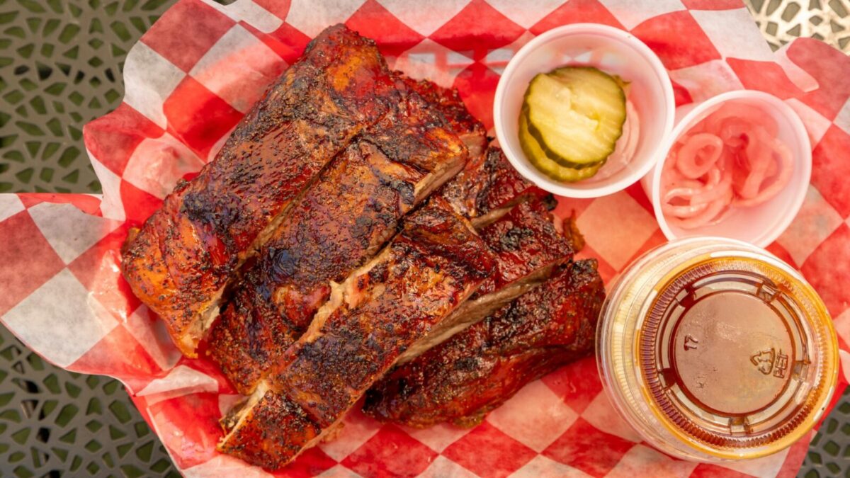 Close-up of grilled barbecue ribs with pickles, onions, and sauce on picnic table.