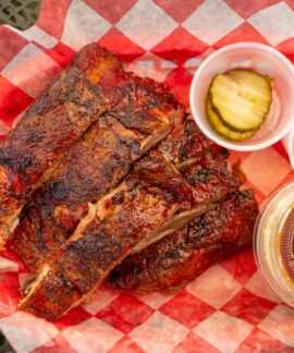 Close-up of grilled barbecue ribs with pickles, onions, and sauce on picnic table.