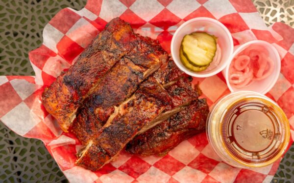 Close-up of grilled barbecue ribs with pickles, onions, and sauce on picnic table.
