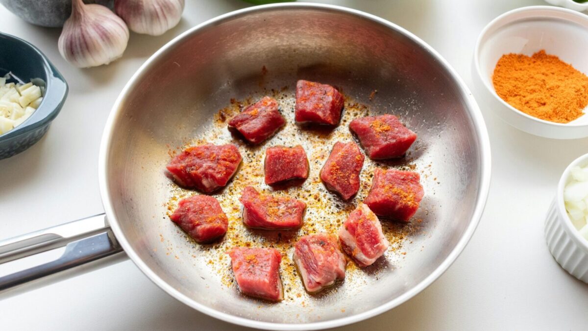 Chunks of raw meat spiced and seared in a skillet surrounded by fresh ingredients.