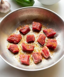 Chunks of raw meat spiced and seared in a skillet surrounded by fresh ingredients.