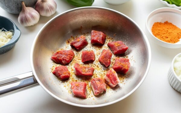 Chunks of raw meat spiced and seared in a skillet surrounded by fresh ingredients.