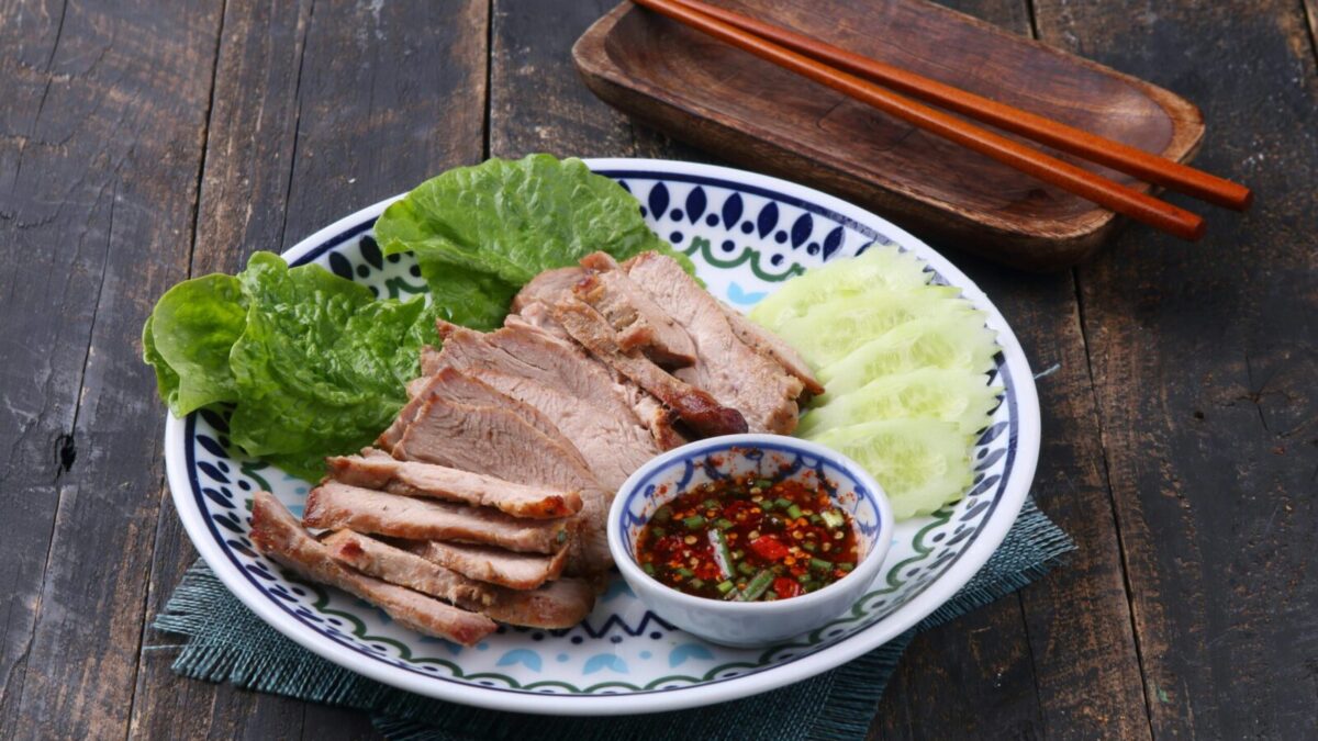 Grilled pork slices with spicy dipping sauce, cucumbers, and lettuce on a decorative plate.