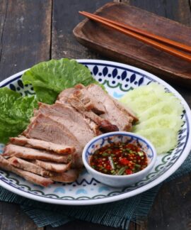 Grilled pork slices with spicy dipping sauce, cucumbers, and lettuce on a decorative plate.