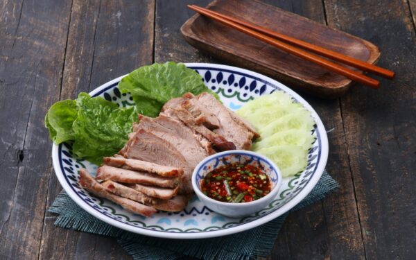 Grilled pork slices with spicy dipping sauce, cucumbers, and lettuce on a decorative plate.
