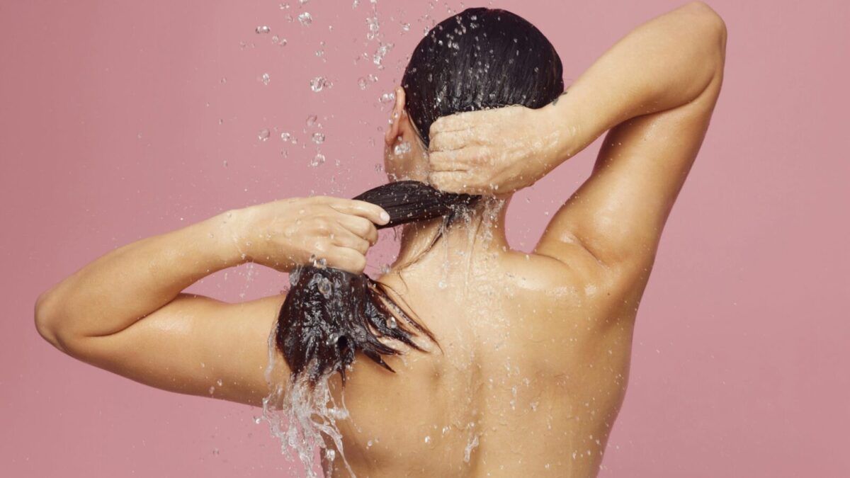 Woman washing long hair under shower with water droplets against a pink background.
