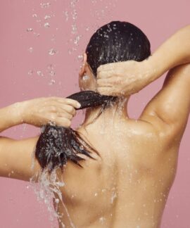 Woman washing long hair under shower with water droplets against a pink background.