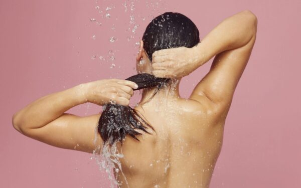 Woman washing long hair under shower with water droplets against a pink background.