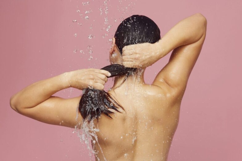 Woman washing long hair under shower with water droplets against a pink background.