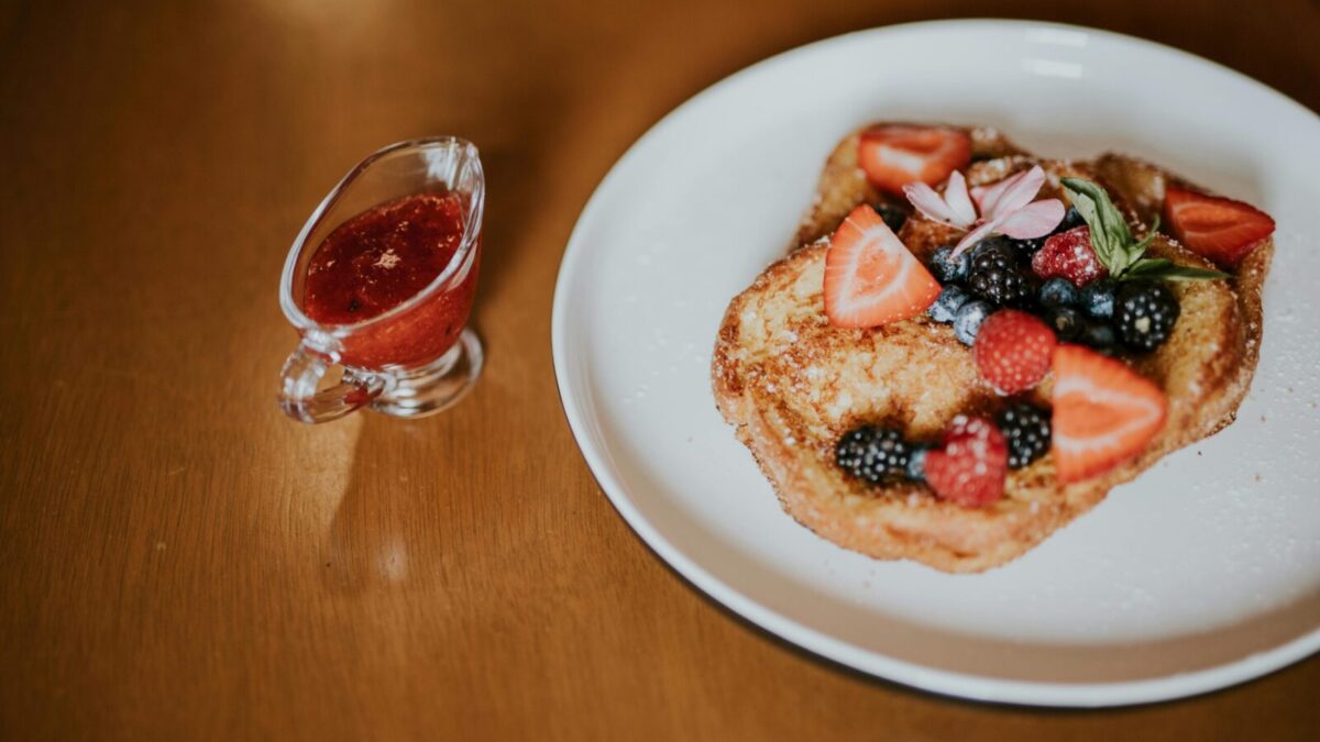Tasty French toast topped with fresh berries and served with a side of berry syrup on a wooden table.