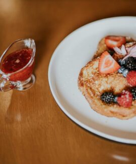 Tasty French toast topped with fresh berries and served with a side of berry syrup on a wooden table.