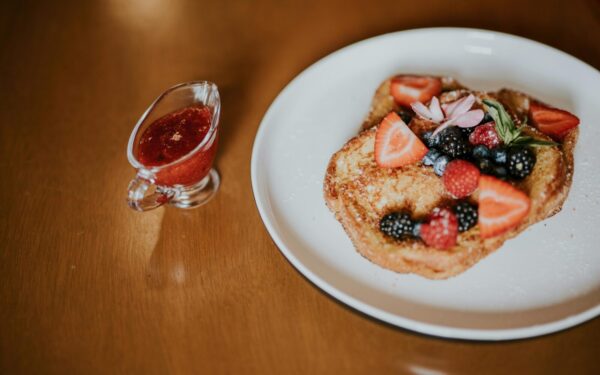 Tasty French toast topped with fresh berries and served with a side of berry syrup on a wooden table.