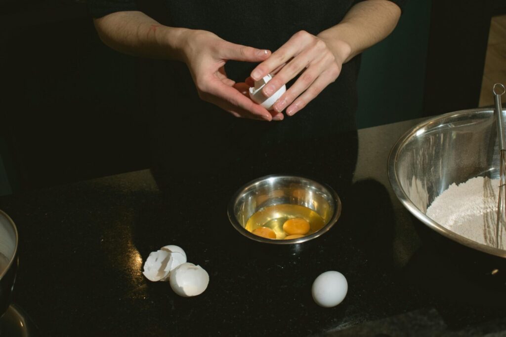 Hands breaking eggs into a metal bowl beside flour ingredients on a dark countertop.