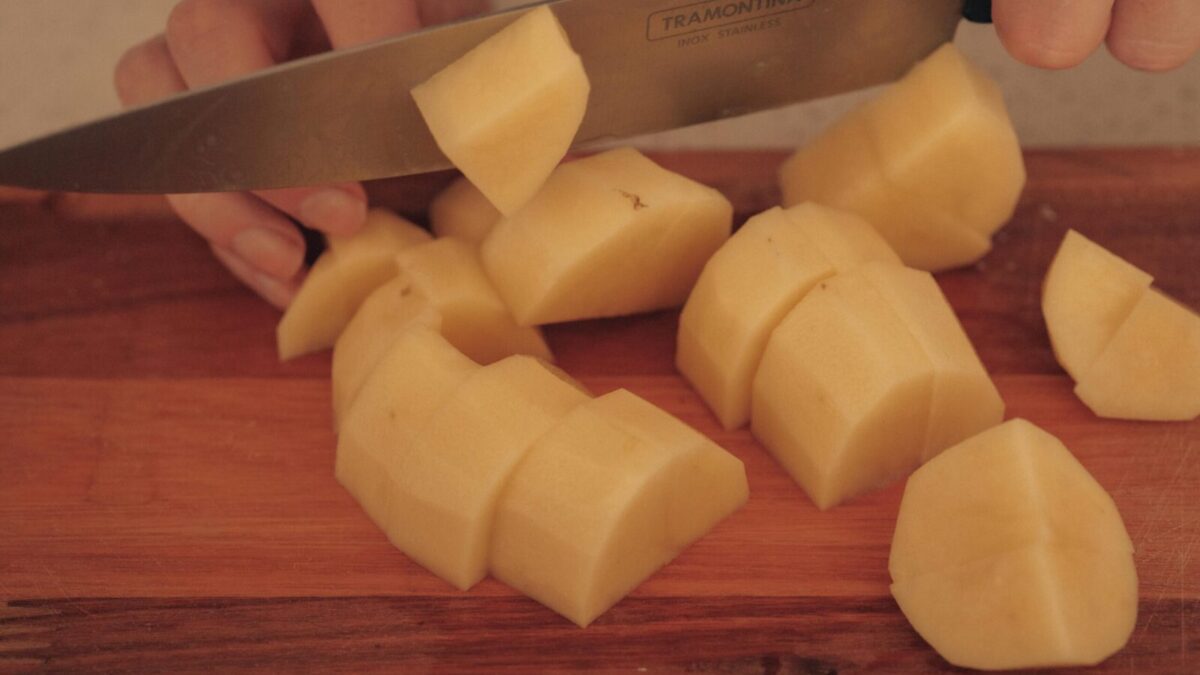 A person cuts potatoes on a wooden board with a kitchen knife.