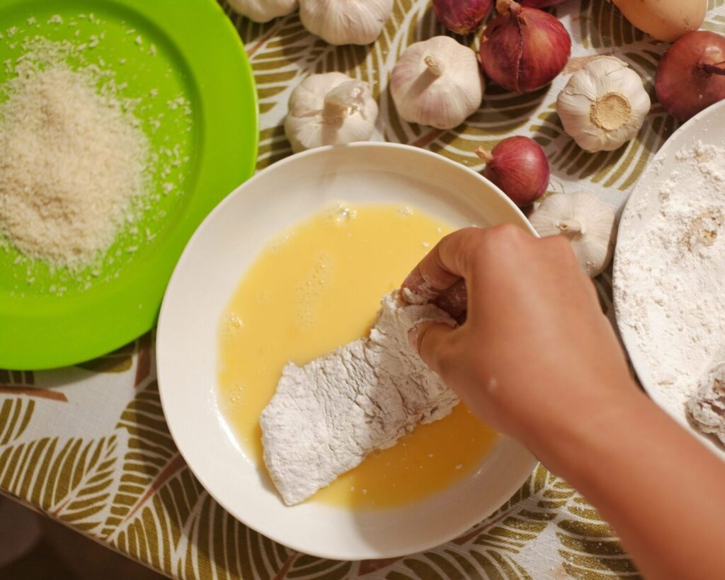 A close-up of a hand dipping flour-coated chicken in egg, surrounded by garlic and onions.