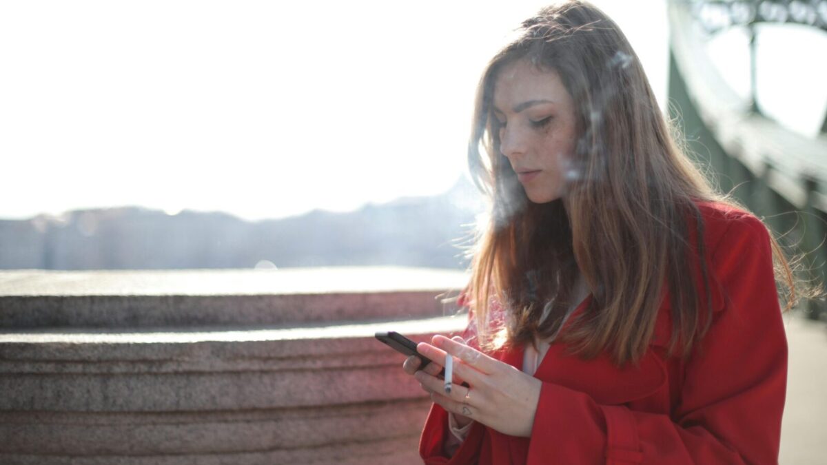 Portrait of a woman in red coat smoking and texting outside on a sunny day.