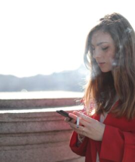 Portrait of a woman in red coat smoking and texting outside on a sunny day.