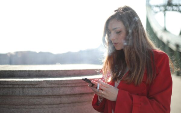 Portrait of a woman in red coat smoking and texting outside on a sunny day.