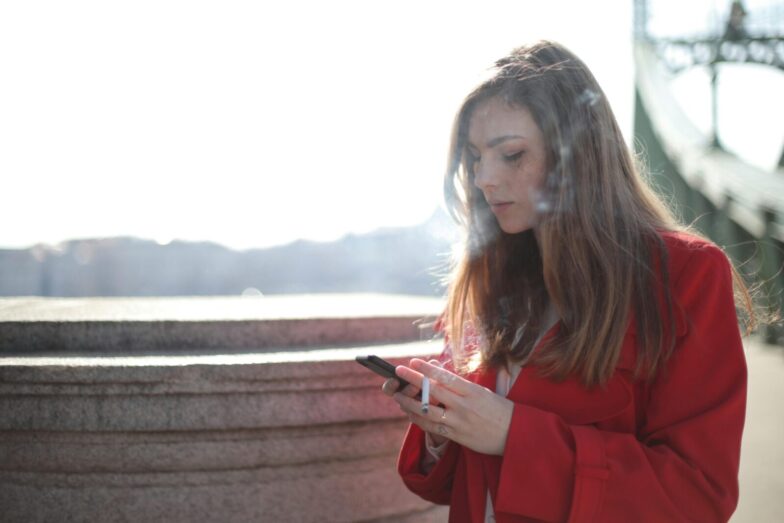 Portrait of a woman in red coat smoking and texting outside on a sunny day.