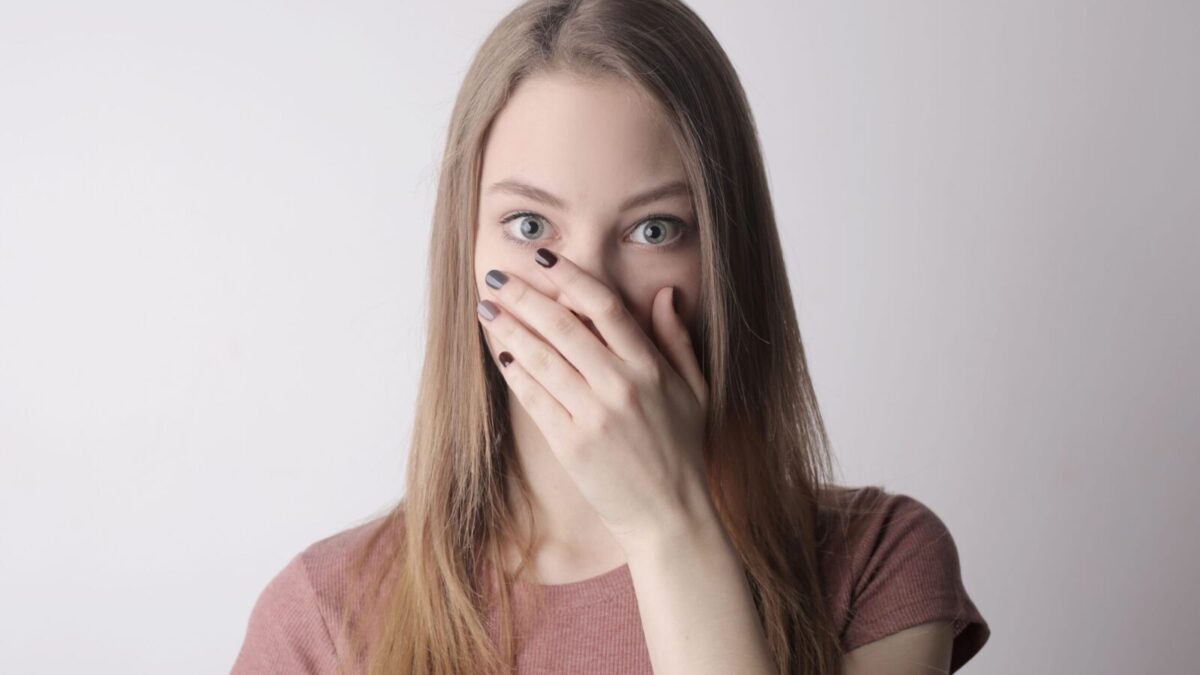 Surprised young woman covering mouth with hand and looking at camera while standing against gray wall background