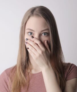 Surprised young woman covering mouth with hand and looking at camera while standing against gray wall background
