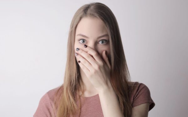 Surprised young woman covering mouth with hand and looking at camera while standing against gray wall background
