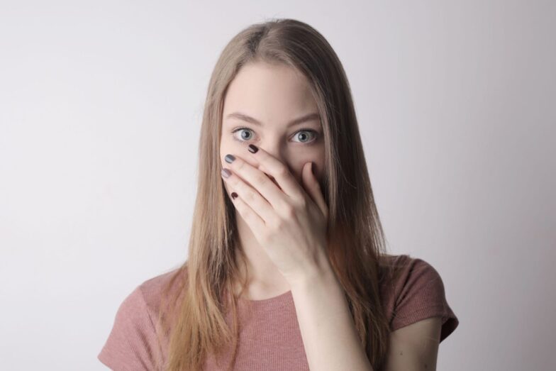 Surprised young woman covering mouth with hand and looking at camera while standing against gray wall background