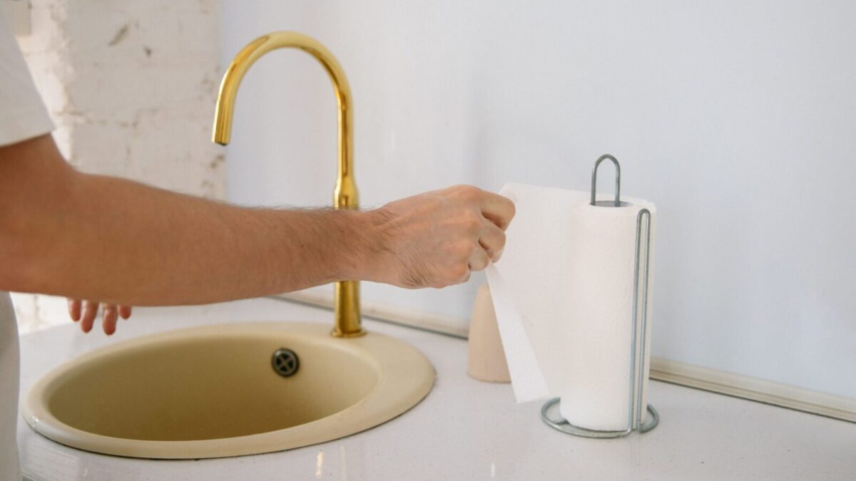 A bathroom sink with a golden faucet and hand reaching for a paper towel in a clean setting.