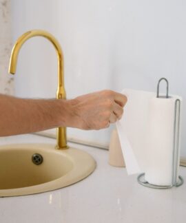 A bathroom sink with a golden faucet and hand reaching for a paper towel in a clean setting.