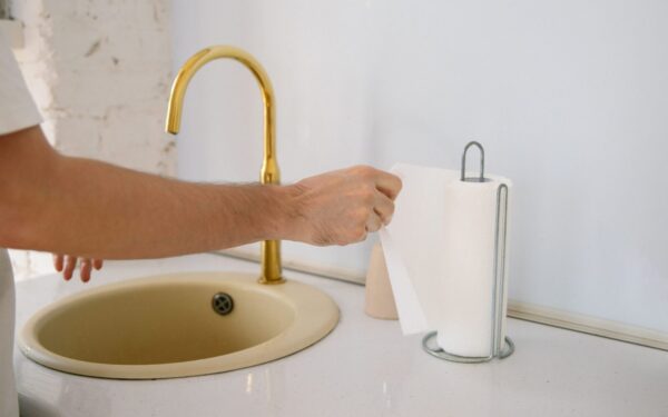 A bathroom sink with a golden faucet and hand reaching for a paper towel in a clean setting.