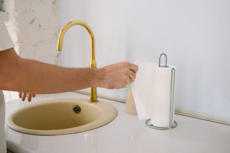 A bathroom sink with a golden faucet and hand reaching for a paper towel in a clean setting.