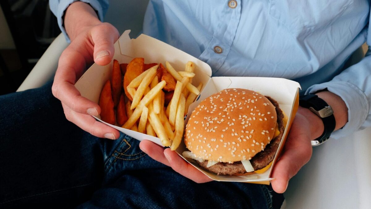 Unrecognizable male with wristwatch in casual clothes sitting on chair and holding box of burger and french fries in hands