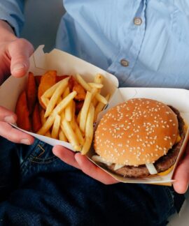 Unrecognizable male with wristwatch in casual clothes sitting on chair and holding box of burger and french fries in hands