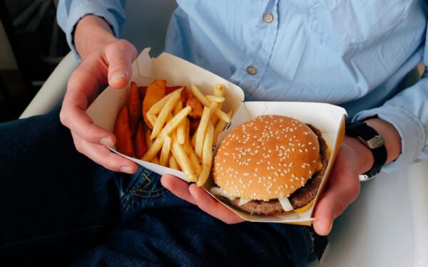 Unrecognizable male with wristwatch in casual clothes sitting on chair and holding box of burger and french fries in hands
