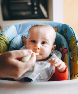 Crop person feeding baby with bib sitting in feeding chair and looking at camera