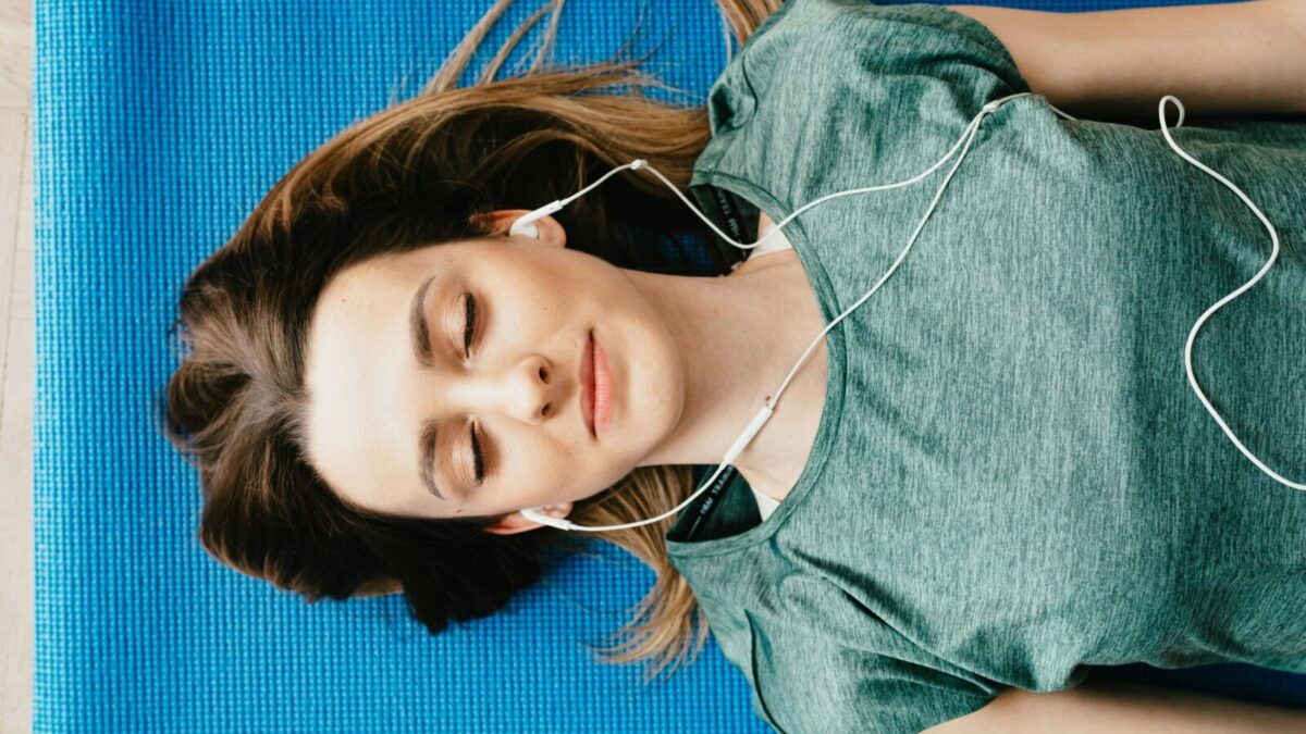From above of calm young female in casual wear listening to music in earphones while taking break after workout on blue mat in flat