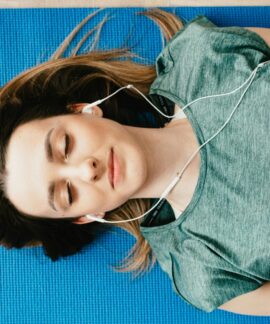 From above of calm young female in casual wear listening to music in earphones while taking break after workout on blue mat in flat