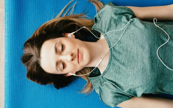 From above of calm young female in casual wear listening to music in earphones while taking break after workout on blue mat in flat