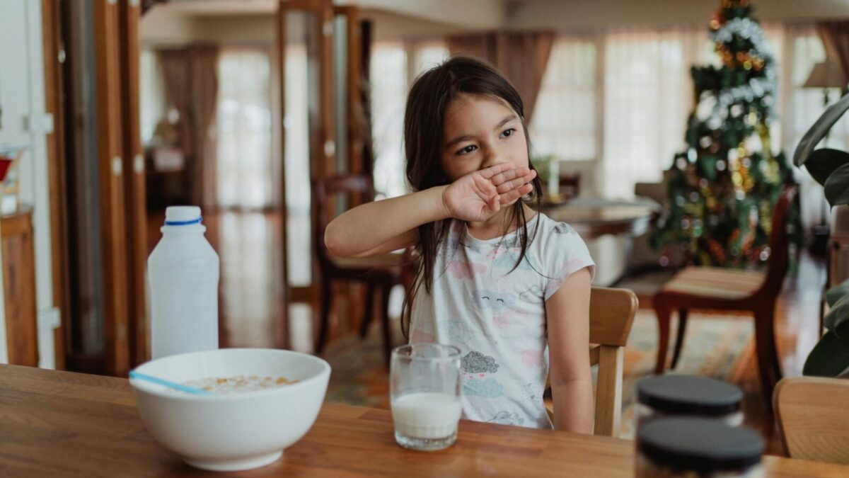 A young girl sits at a wooden table enjoying cereal and milk in a cozy home setting.