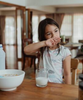 A young girl sits at a wooden table enjoying cereal and milk in a cozy home setting.