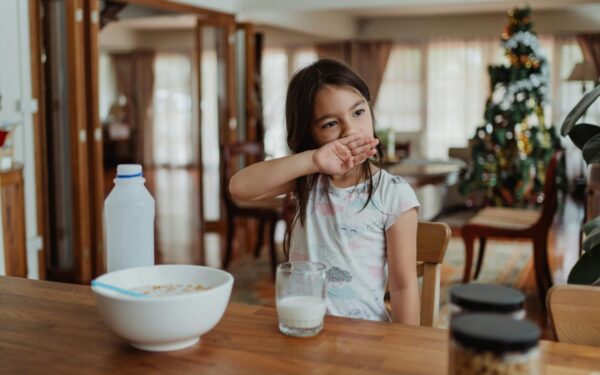A young girl sits at a wooden table enjoying cereal and milk in a cozy home setting.