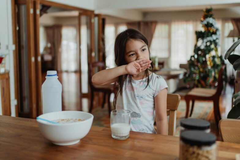 A young girl sits at a wooden table enjoying cereal and milk in a cozy home setting.