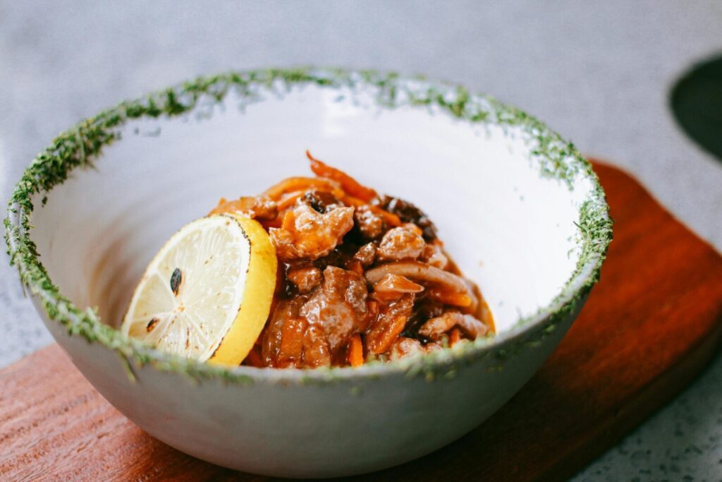 A flavorful chicken dish in a ceramic bowl with a lemon slice and herb garnish on a wooden board.