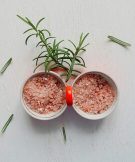 Top view of pink Himalayan salt and fresh rosemary in bowls on white table.