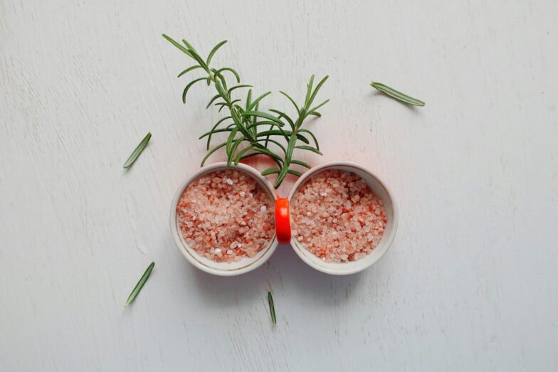 Top view of pink Himalayan salt and fresh rosemary in bowls on white table.
