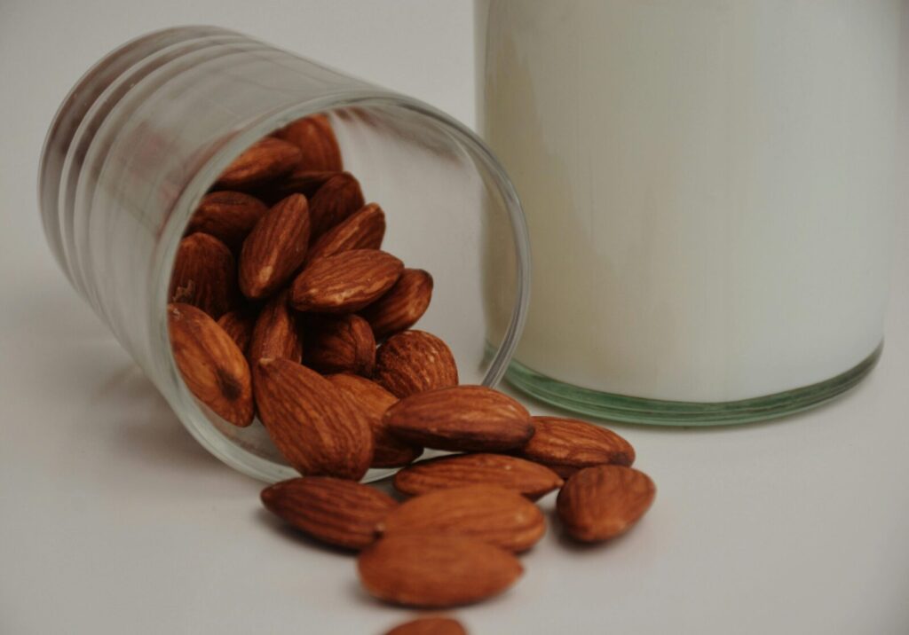 Transparent glass with spilled whole crispy almonds near dairy beverage on white background