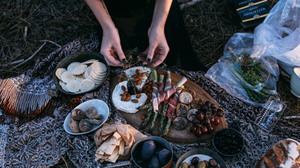 From above crop anonymous female serving yummy food including asparagus with bacon potatoes and grapes on wooden board placed on plaid during picnic in nature