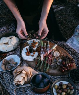 From above crop anonymous female serving yummy food including asparagus with bacon potatoes and grapes on wooden board placed on plaid during picnic in nature