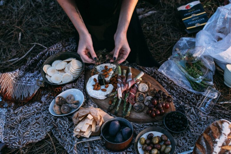 From above crop anonymous female serving yummy food including asparagus with bacon potatoes and grapes on wooden board placed on plaid during picnic in nature