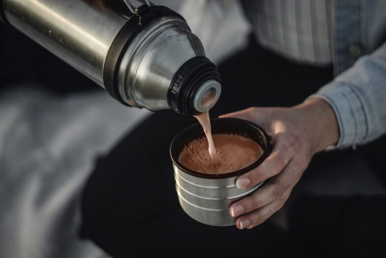 From above of crop anonymous female pouring aromatic hot coffee in thermos cap on blurred background